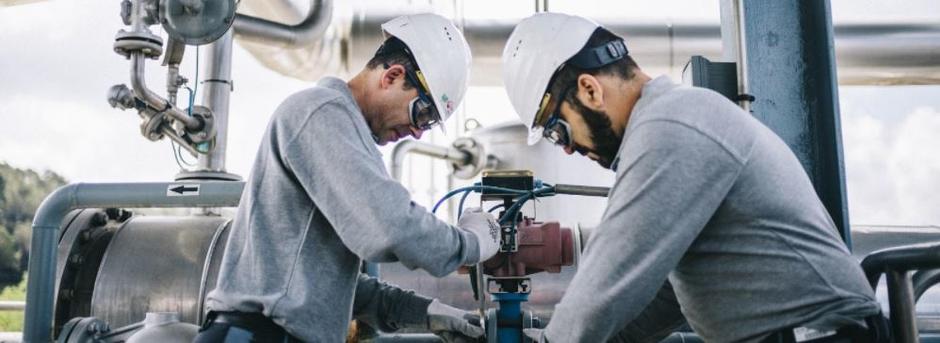 Zwei Arbeiter, welche gemeinsam an einer Pumpe arbeiten. Sie tragen weiße Schutzhelme, Schutzbrillen und graue Kleidung.
