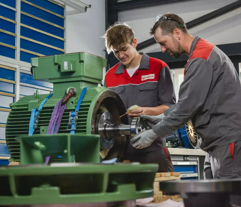 Ein erfahrener Mechaniker und ein jüngerer Kollege arbeiten gemeinsam an einem großen grünen Industriemotor oder einer Pumpe in einer Werkstatt. Beide tragen graue und rote Arbeitsuniformen mit dem „VEOLIA“-Logo. Der ältere Mechaniker, der eine Schutzbrille trägt, hält ein Lagerteil und schaut auf die Arbeit, während der jüngere Kollege, der lächelt, Öl auf eine Welle oder ein Lager aufträgt. Im Vordergrund ist unscharf ein weiteres grünes Maschinenteil zu sehen. Der Hintergrund zeigt blaue Regale und eine 
