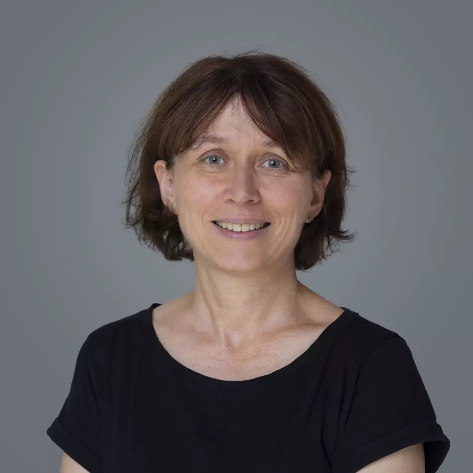 Portraitfoto einer lächelnden Frau mittleren Alters mit kurzen braunen Haaren, die ein schwarzes T-Shirt trägt vor grauem Hintergrund.
