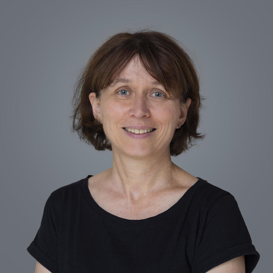Portraitfoto einer lächelnden Frau mittleren Alters mit kurzen braunen Haaren, die ein schwarzes T-Shirt trägt vor grauem Hintergrund.